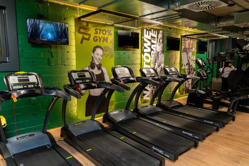 Wnętrze siłowni 100%GYM w Chojnicach, strefa cardio z logo marki na ścianie